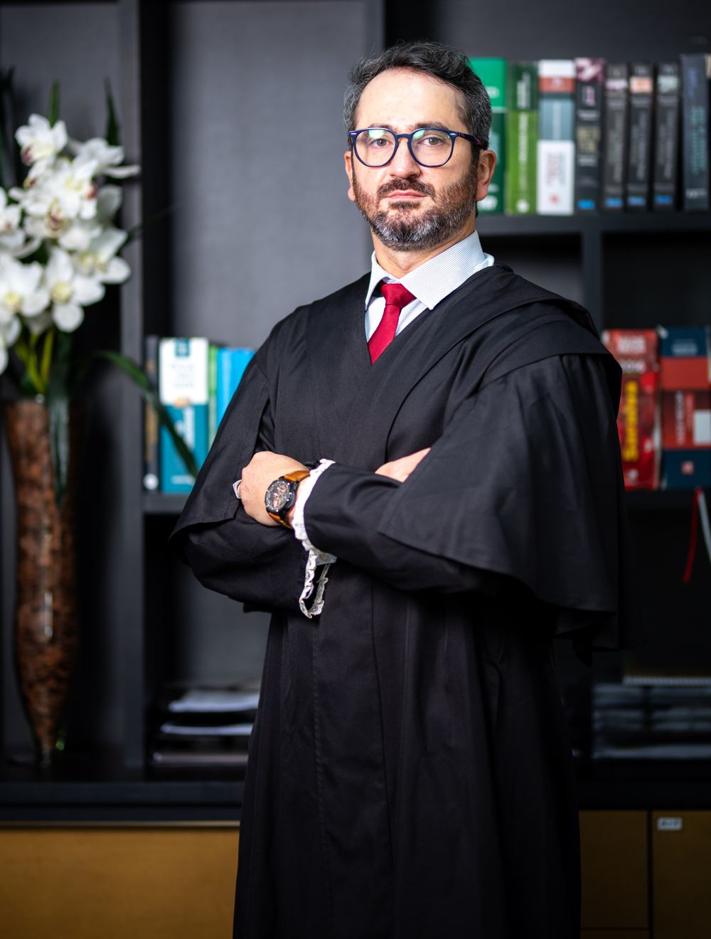 Roger Ellwanger de beca, braços cruzados, diante da estante de livros jurídicos